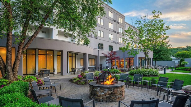 An outdoor patio with stone fire pit seating, surrounded by lush greenery and a modern building with large windows.
