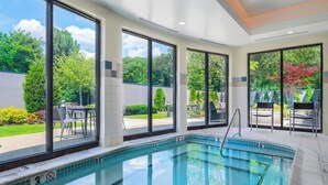 A swimming pool with clear blue water, surrounded by large windows offering views of greenery and a patio area with outdoor furniture.