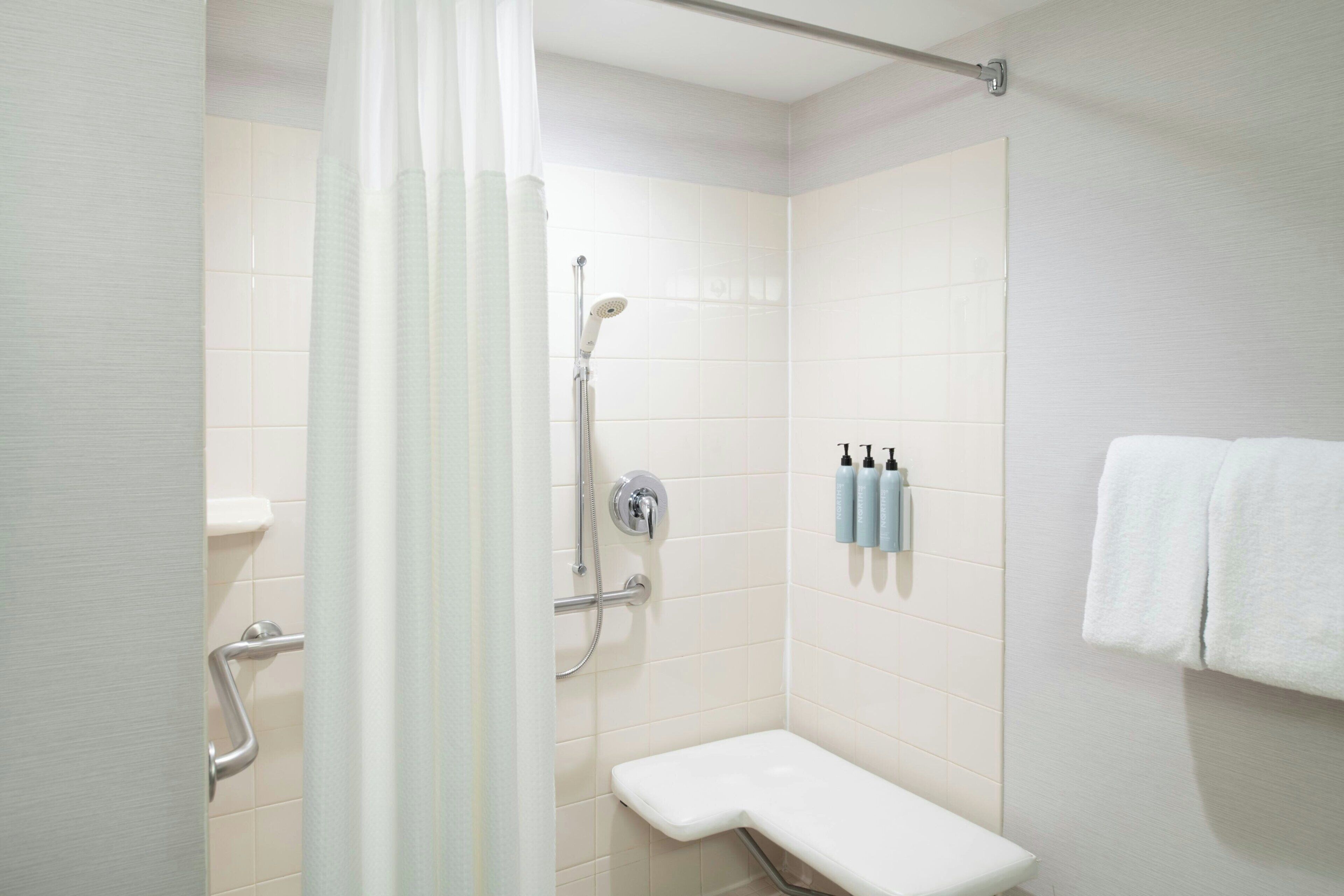 A small, modern bathroom with white tiles, a shower with a handheld showerhead, a bench, and a towel rack with towels.