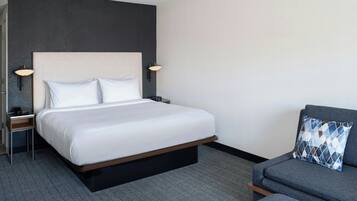 Een hotelkamer met een groot bed, twee nachtkastjes met lampen, een grijze bank en een kamerkleed met dessin.