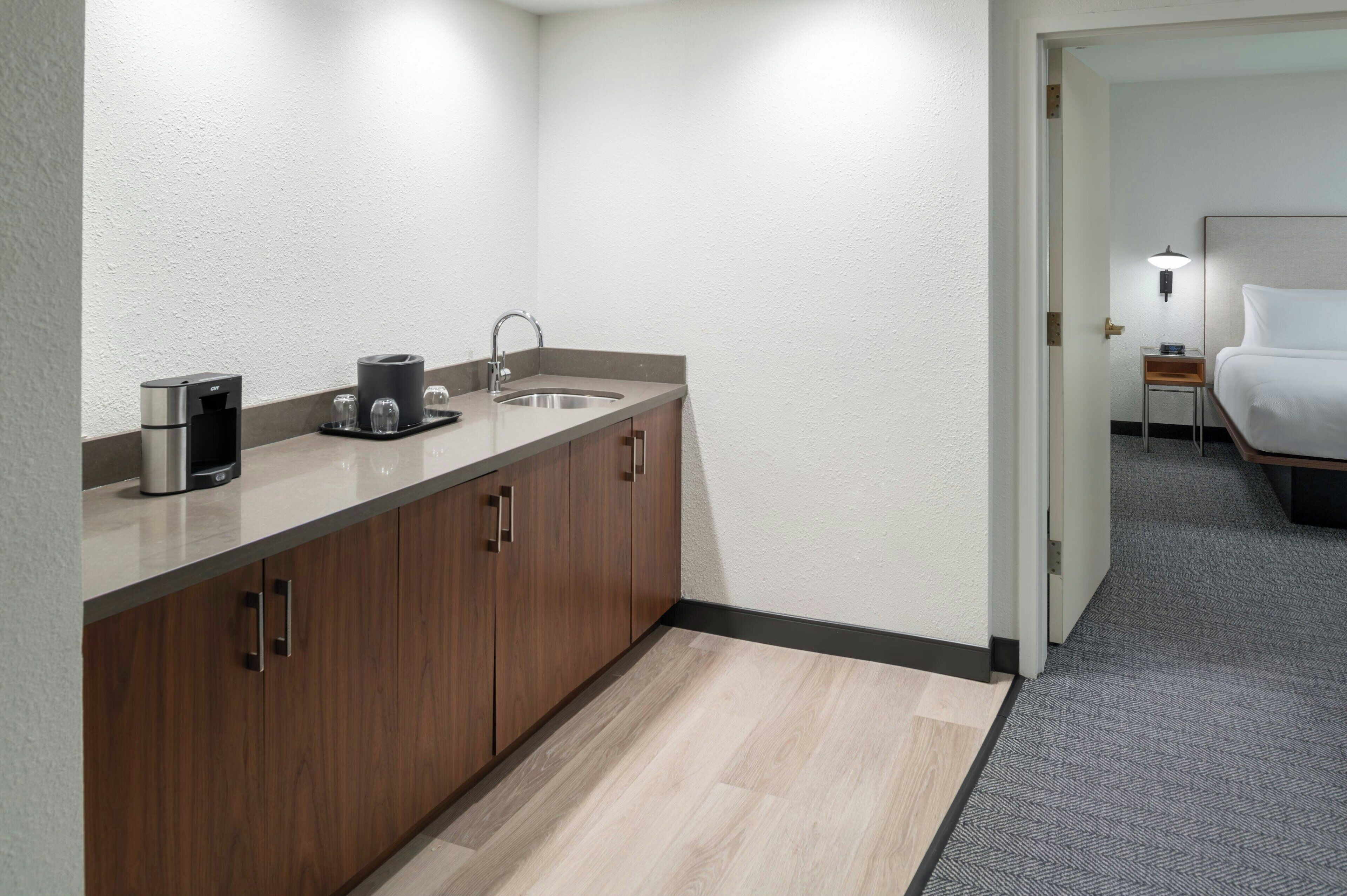 Una habitación de hotel moderna con kitchenette que incluye cafetera, lavamanos y armarios de madera.
