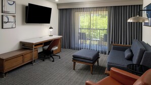 Ett modernt hotellrum med en platt-TV som är monterad på väggen, ett skrivbord med en stol, en soffa, en pall och ett litet bord.