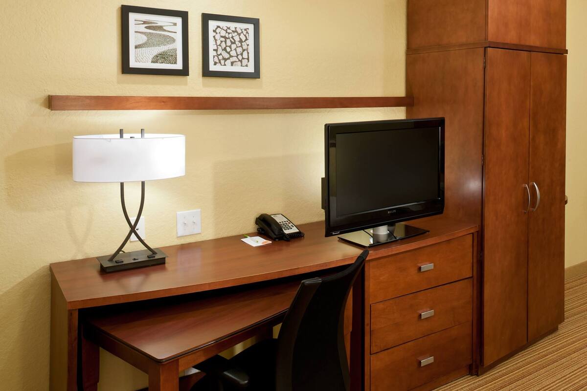 Quarto de hotel com secretária de madeira, cadeira de escritório preta, televisão de tela plana, luminária, telefone e guarda-roupa.