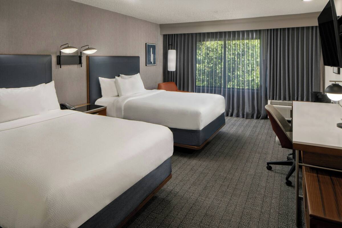 Quarto de hotel com duas camas, uma escrivaninha, uma cadeira, uma televisão e uma janela com cortinas.