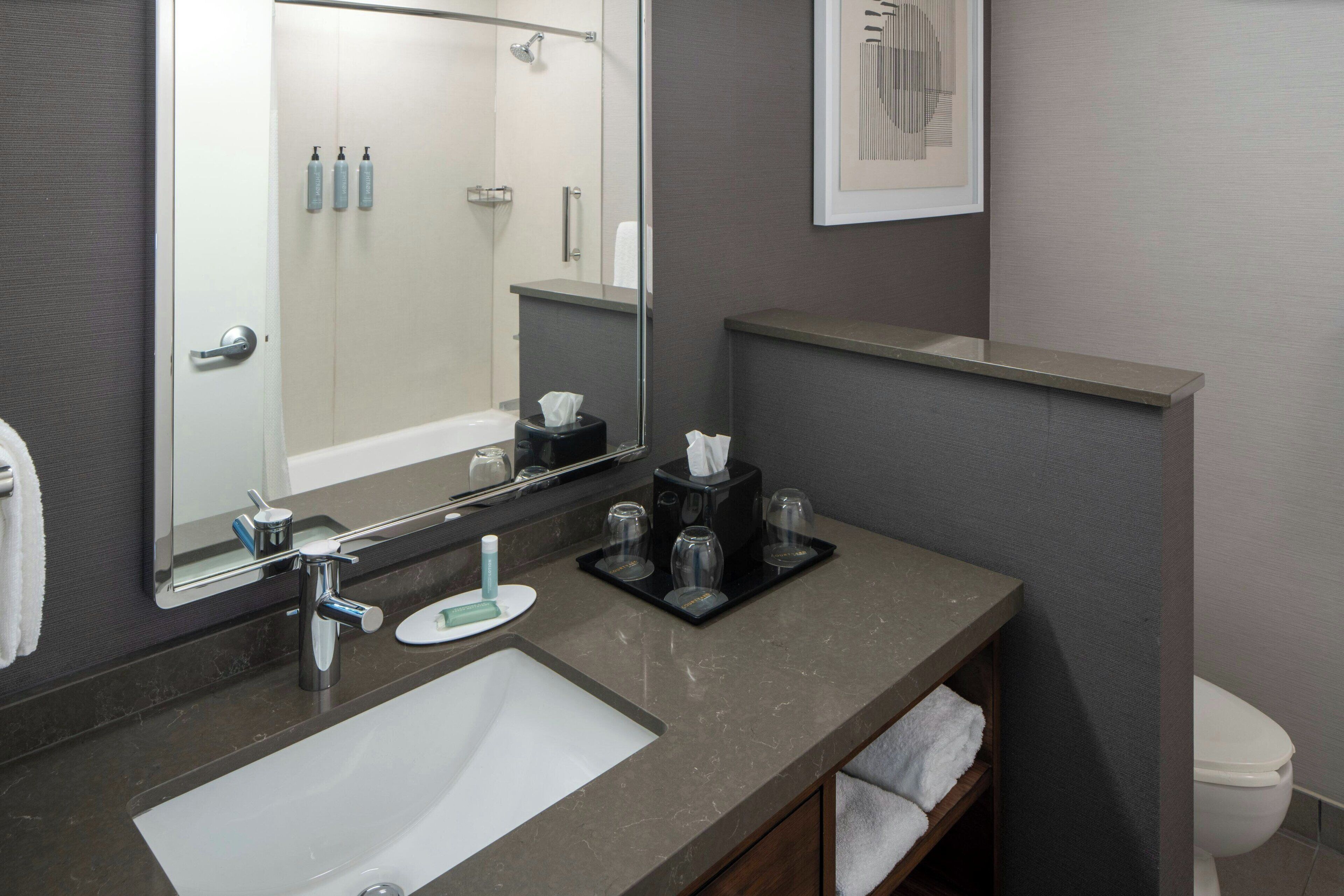 A modern bathroom with a large mirror, a sink, and a toilet. There are toiletries on the counter and a towel rack.
