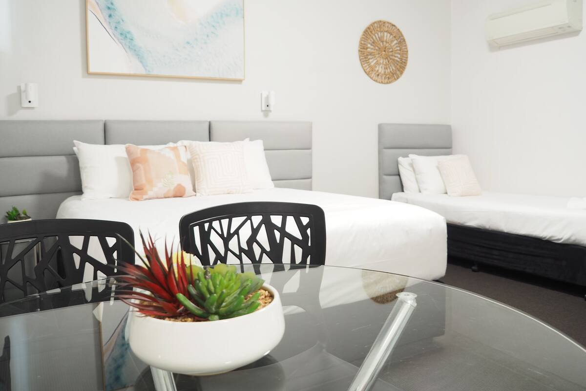 A modern hotel room with a glass dining table, two beds, and a decorative wall piece.