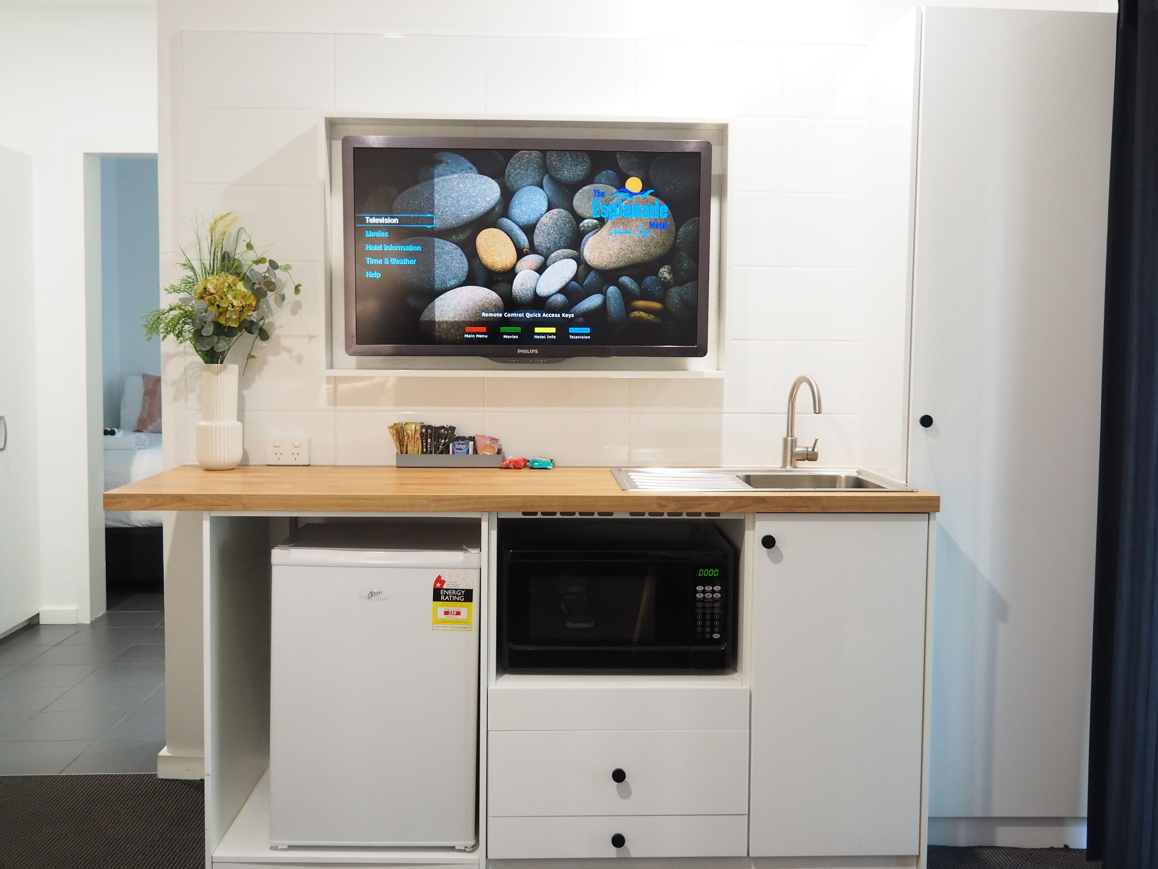A compact kitchenette with a flat-screen TV displaying a travel channel, a small refrigerator, a microwave, and a sink.