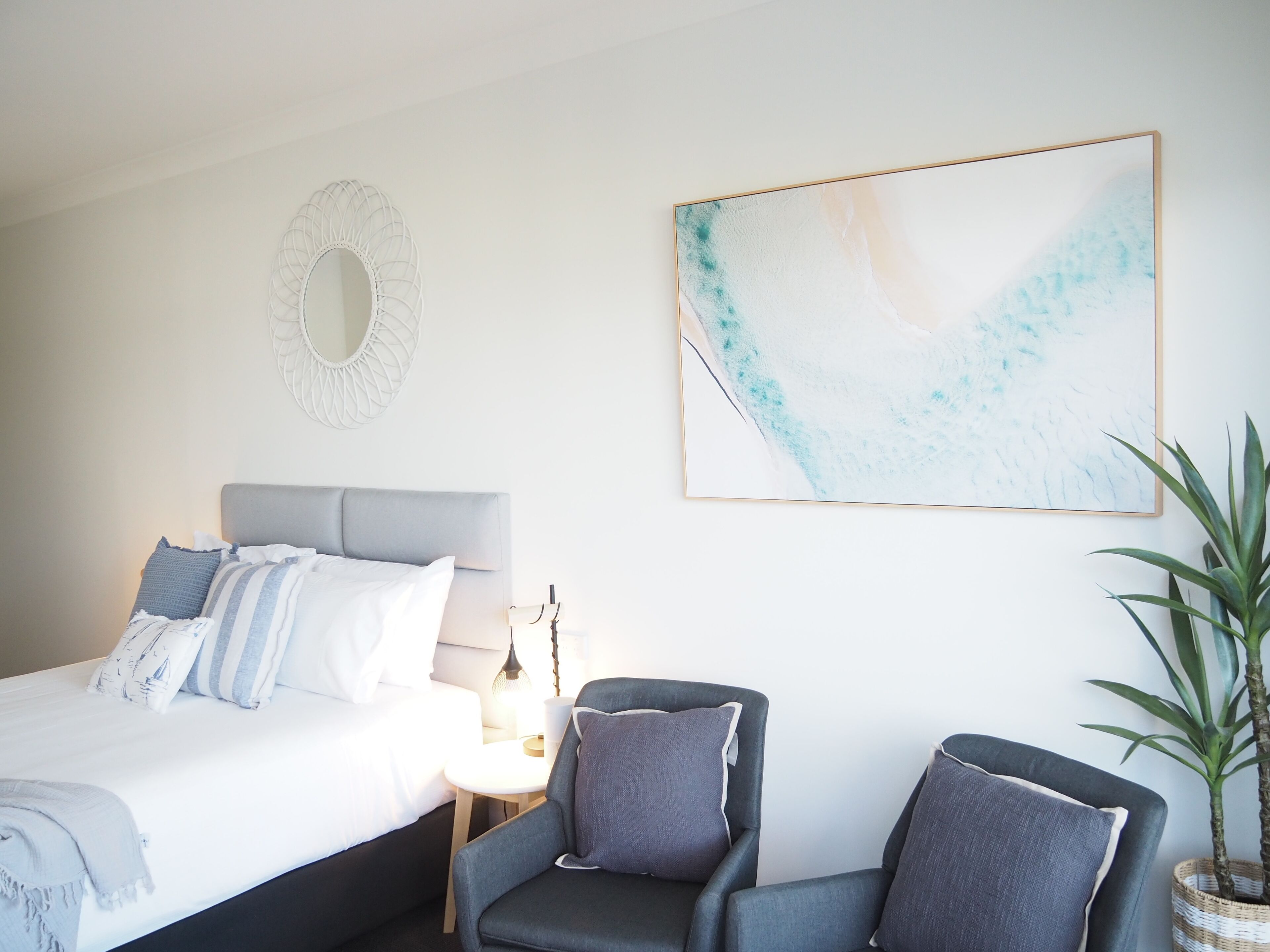Un dormitorio con una cama, dos sillones, una mesita de noche con lámpara, un espejo en la pared y un cuadro abstracto de gran tamaño.