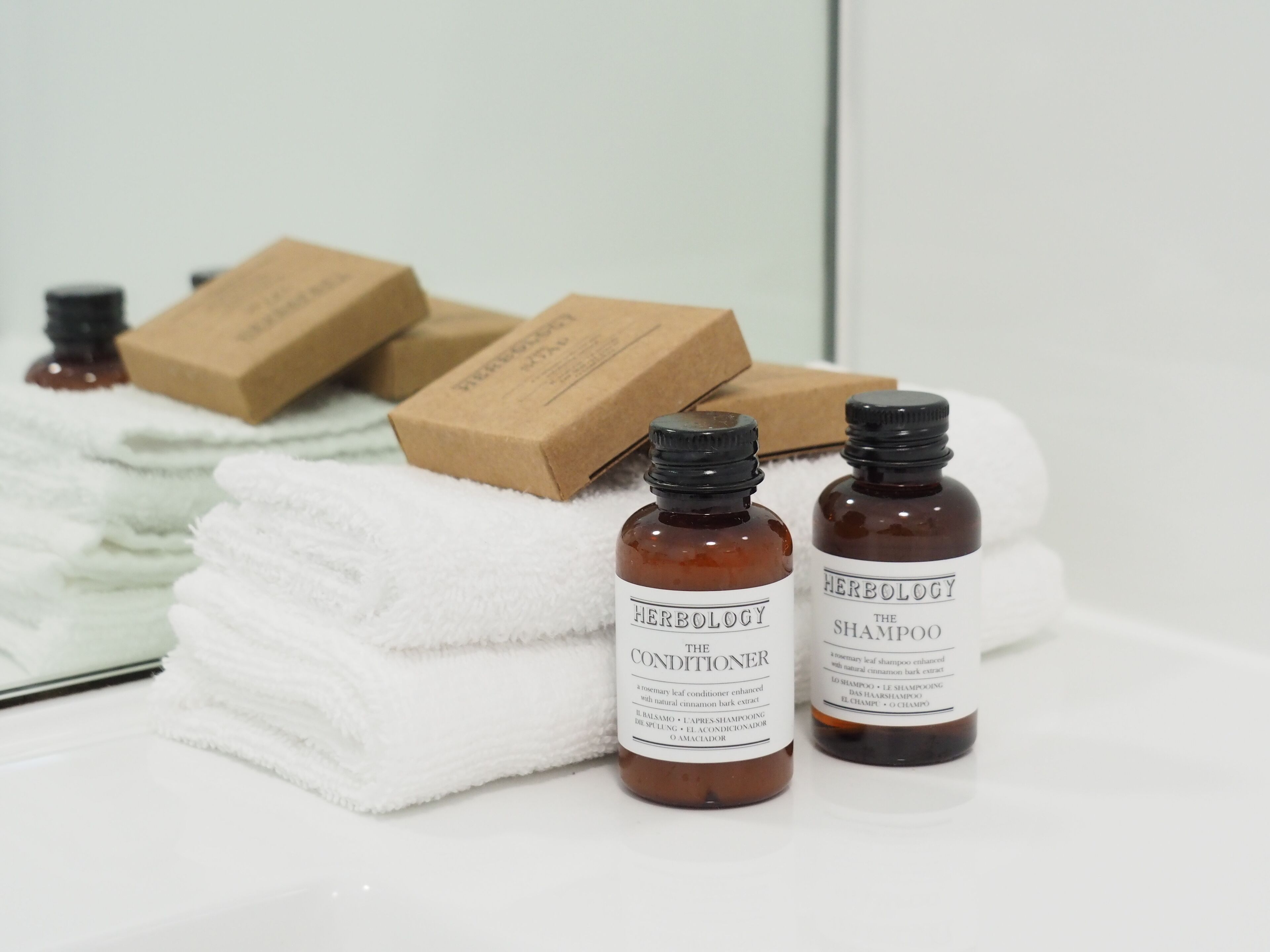 Ein Badezimmer mit weißen Handtüchern, zwei braune Flaschen mit dem Aufdruck 'Herbology The Conditioner' und 'Herbology The Shampoo' sowie einigen Kartons.