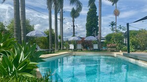 A swimming pool surrounded by palm trees and umbrellas, with a clear sky. - The Esplanade Motel (Warners Bay)