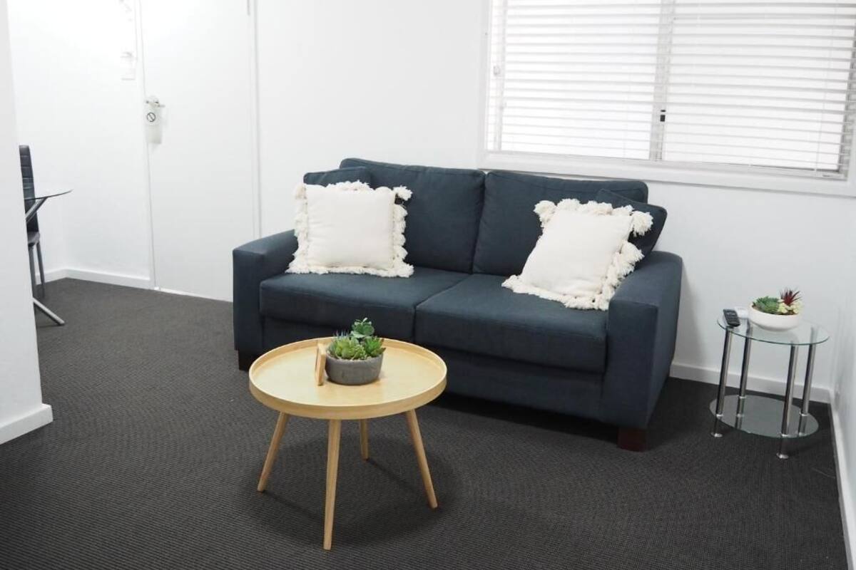 A room with a dark blue sofa, two white pillows, a small round wooden table with a plant, and a glass side table with a plant.