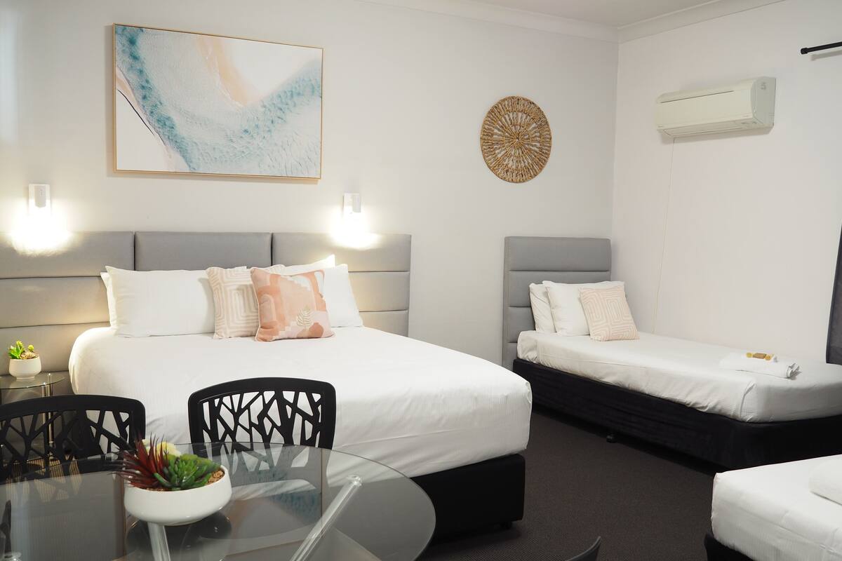 A hotel room with two beds, a glass table with chairs, a wall-mounted air conditioner, and a decorative wall piece.
