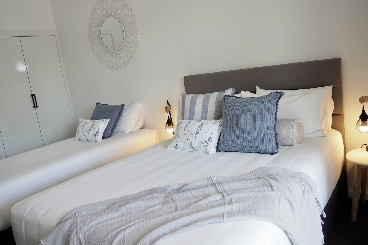 A double bed with white linens, a grey throw blanket, and multiple pillows in a modern bedroom with a grey headboard, bedside tables with lamps, and a white wardrobe.