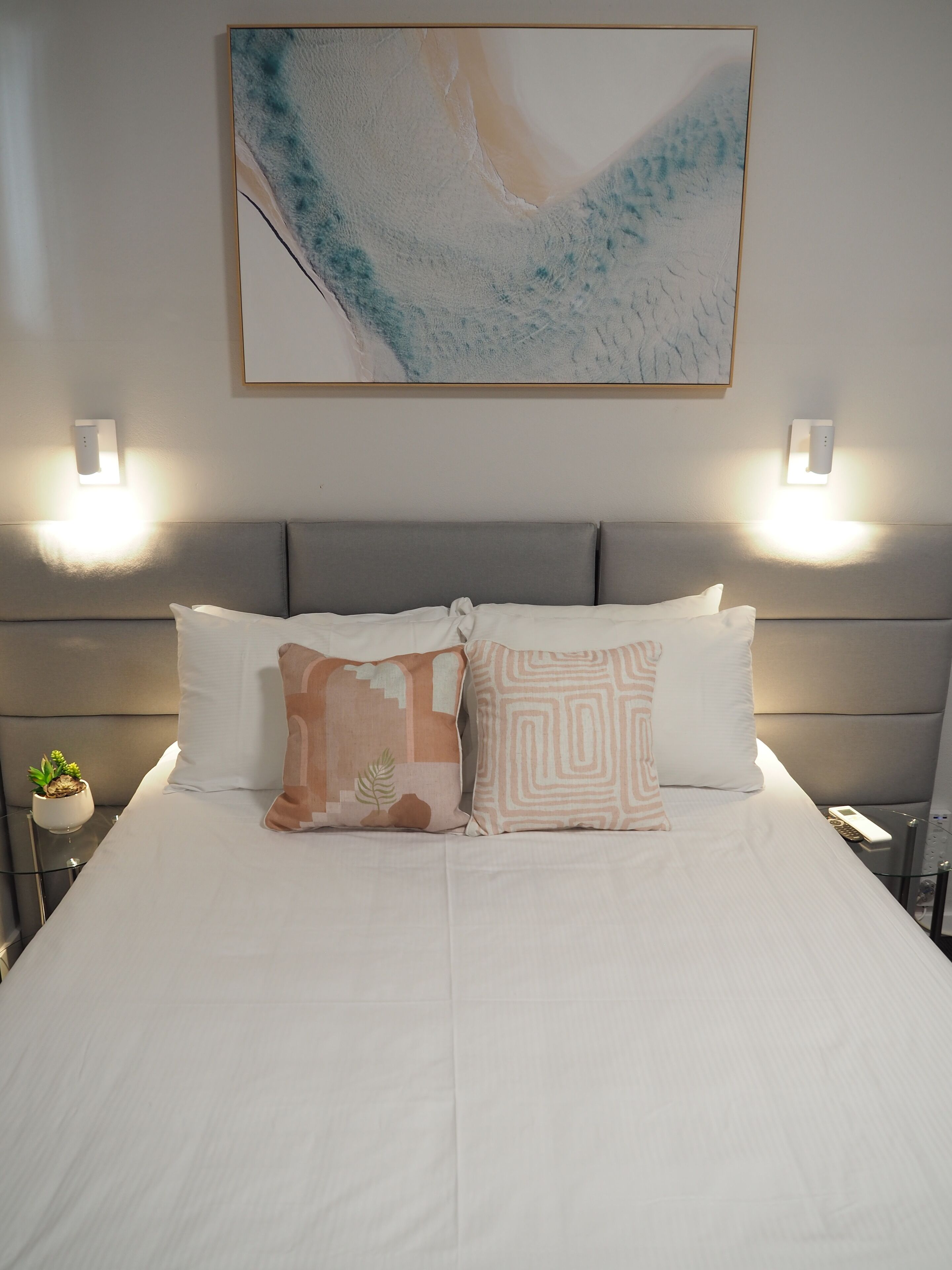 A neatly made bed with two pillows, a small plant on a bedside table, and a wall-mounted light fixture.