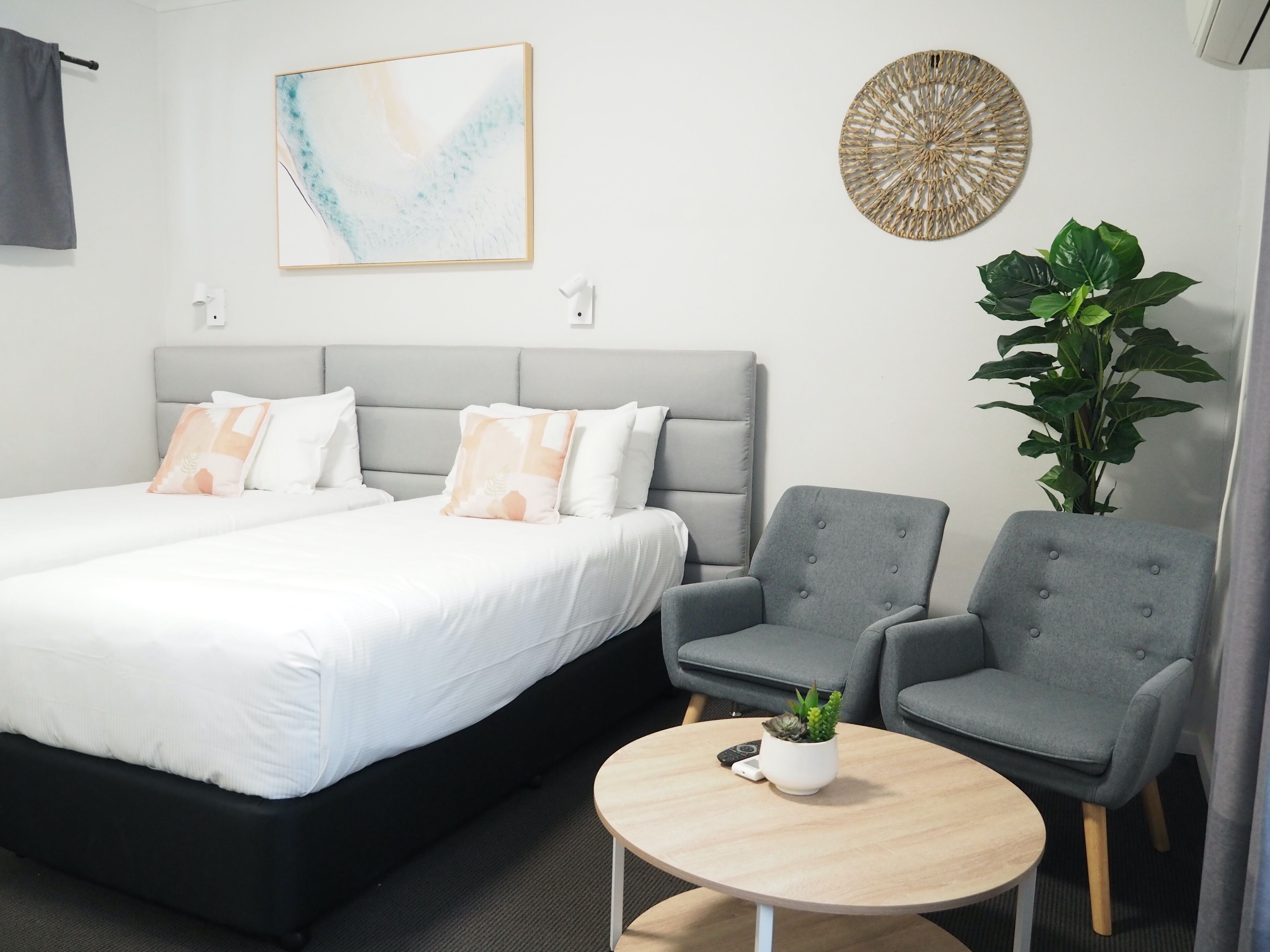 A modern hotel room with a large bed, two grey armchairs, a small round table with a plant, a wall-mounted artwork, and a woven decorative piece.