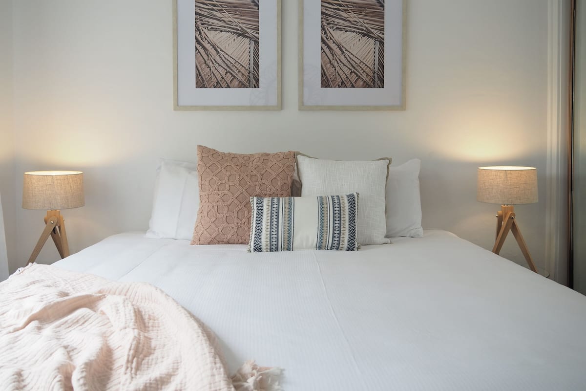 A neatly made bed with white linens, two patterned throw pillows, and a beige blanket. There are two bedside lamps on wooden stands, and two framed abstract paintings on the wall above the bed.