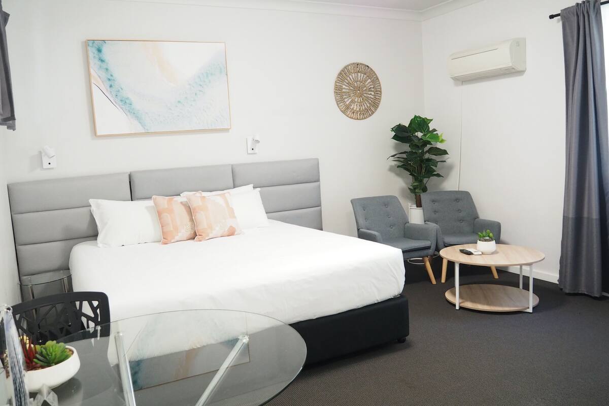 A modern hotel room with a large bed, a small round table, two armchairs, a potted plant, and a wall-mounted air conditioner.