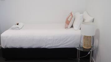Een net opgemaakt bed met witte lakens, één kussen en een opgevouwen handdoek. Een lamp met een geweven kap staat op een glazen bijzettafeltje naast het bed.