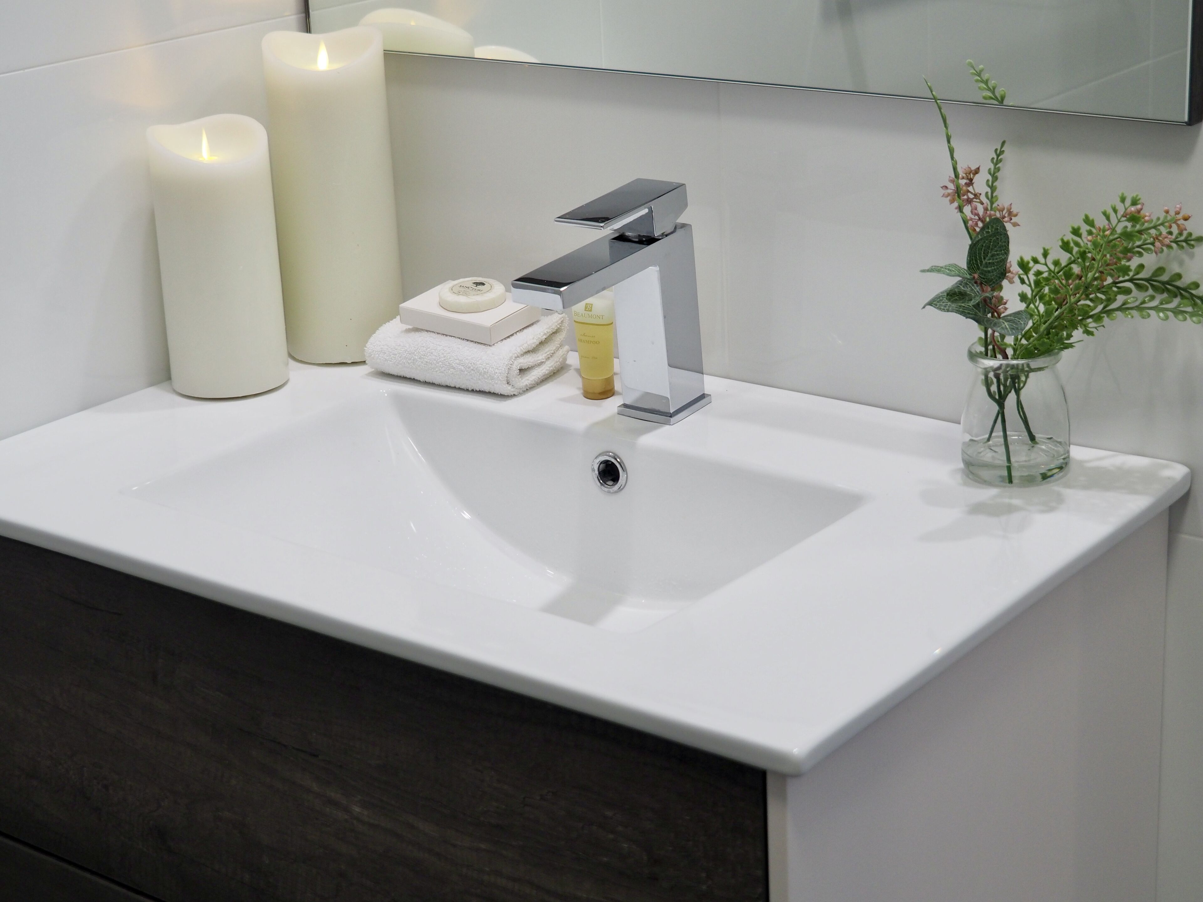 Ein modernes Badezimmer mit Waschbecken in Weiß, einem silbernen Wasserhahn, zwei weißen Kerzen, einem weißen Handtuch, einer kleinen Seifenflasche und einer Vase mit Grünpflanzen.