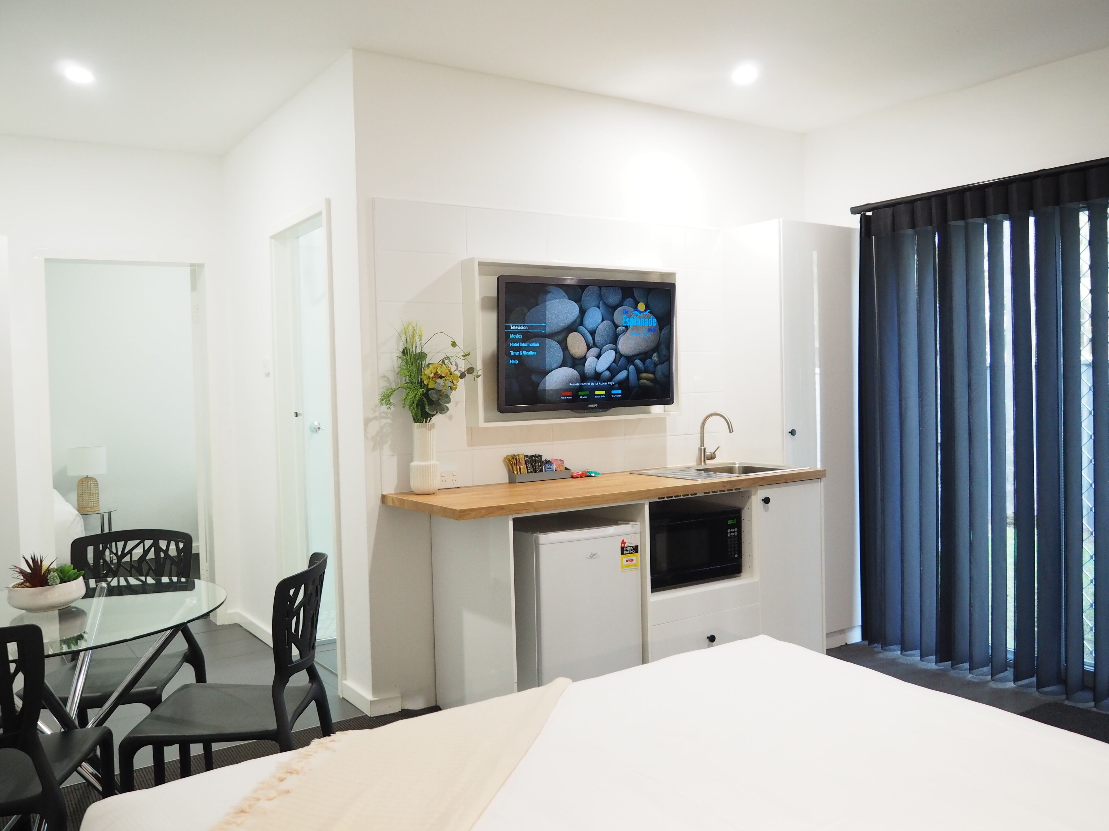 A modern hotel room with a bed, a small kitchenette, a TV mounted on the wall, and a window with curtains.