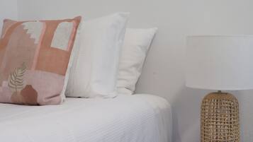 Un letto con biancheria bianca e un cuscino a motivi geometrici, una parete bianca liscia e una lampada in vimini su un tavolino in vetro accanto al letto.