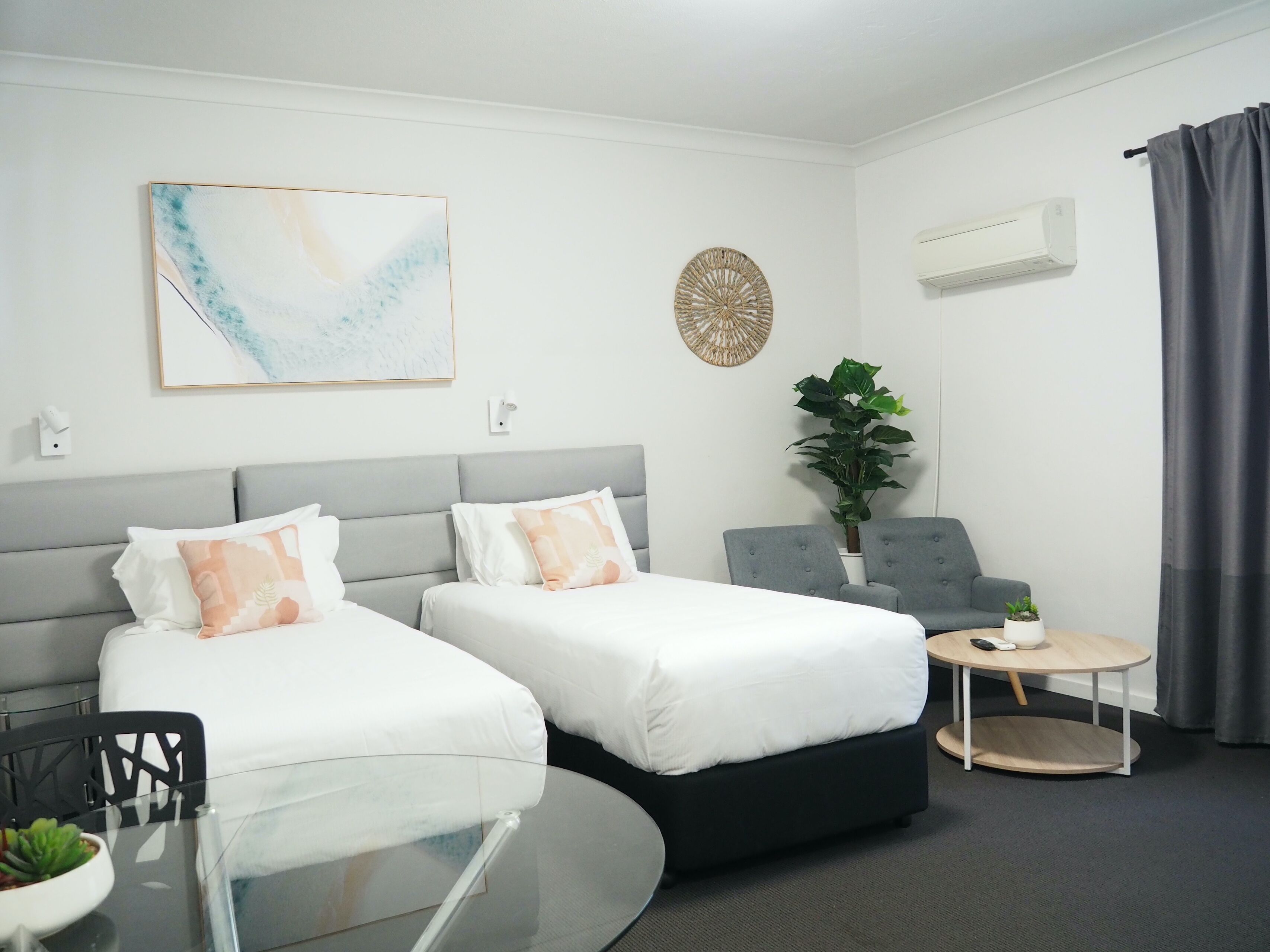 A hotel room with two beds, a small table, a chair, a potted plant, and a wall-mounted air conditioner.