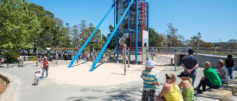 Ein Spielplatz mit einer blauen und roten Kletterkonstruktion, einer Rutsche und einem Sandspielbereich.
