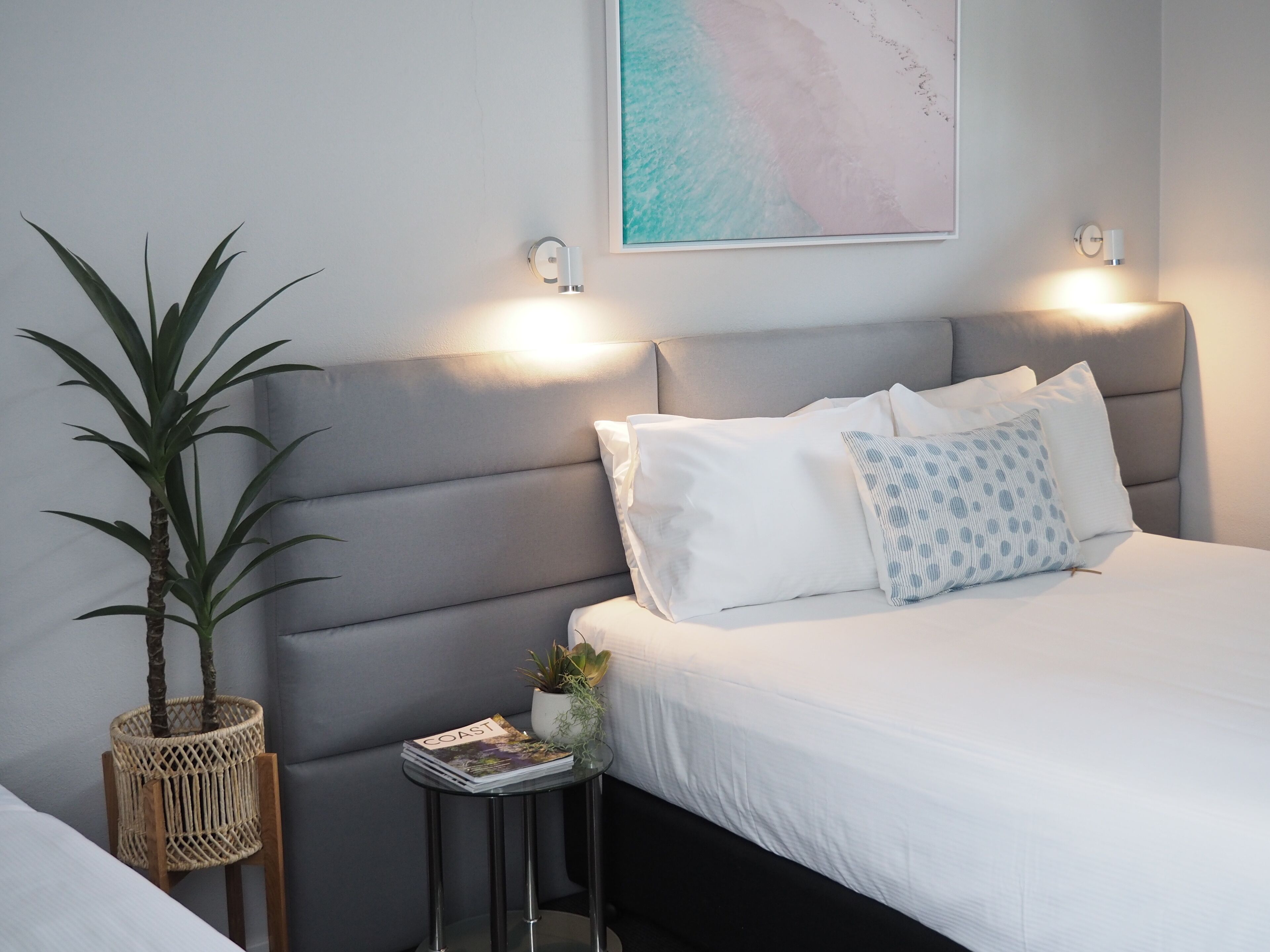 A neatly arranged bedroom with a bed, a bedside table with a magazine, a potted plant, and a wall-mounted light fixture.