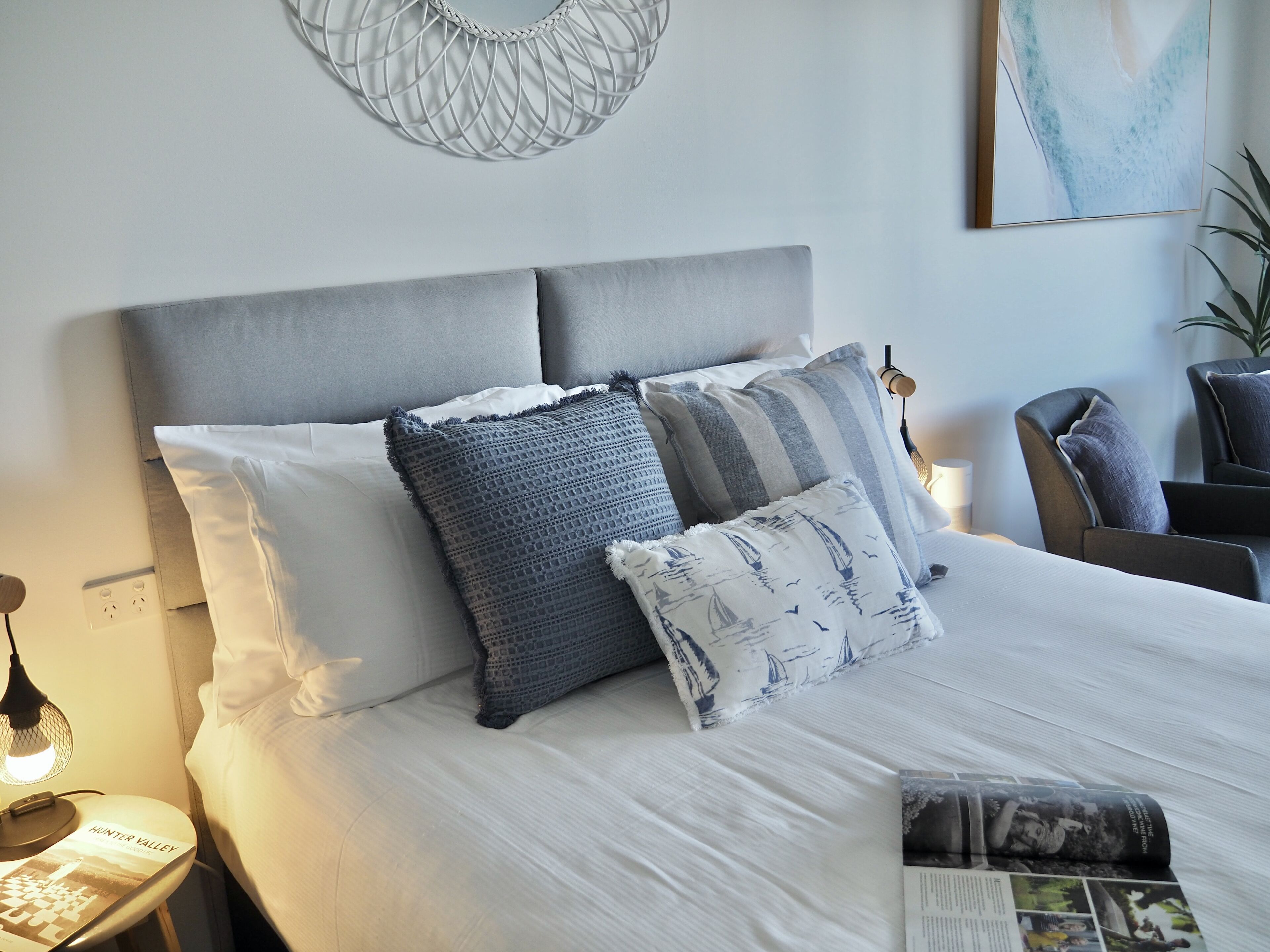 Una cama bien hecha con cabecera gris, varios almohadones, una lámpara de noche y una revista en la mesita.