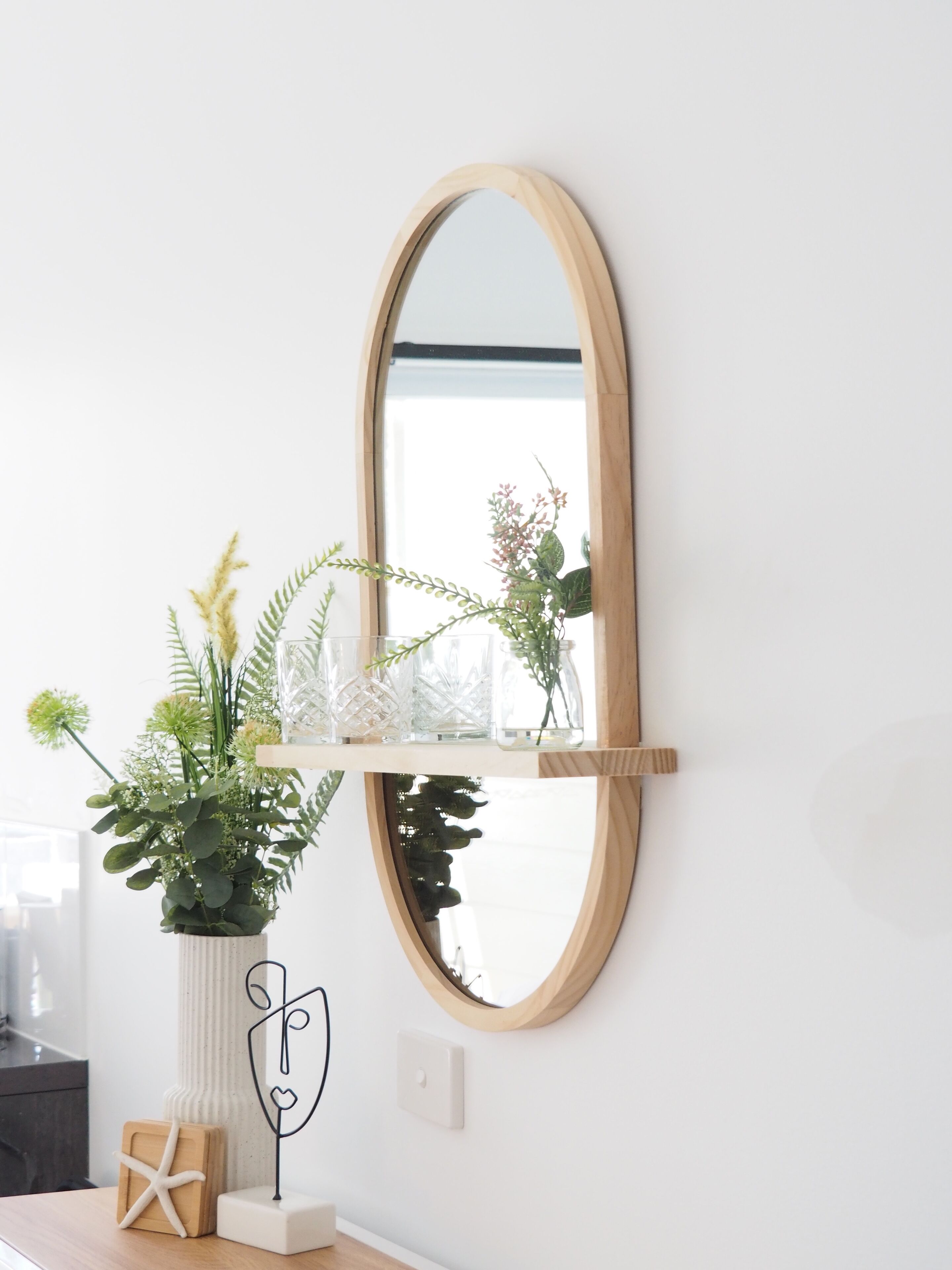 A mirror with a wooden frame mounted on a white wall, a white vase with green plants, and a small white shelf with decorative items.