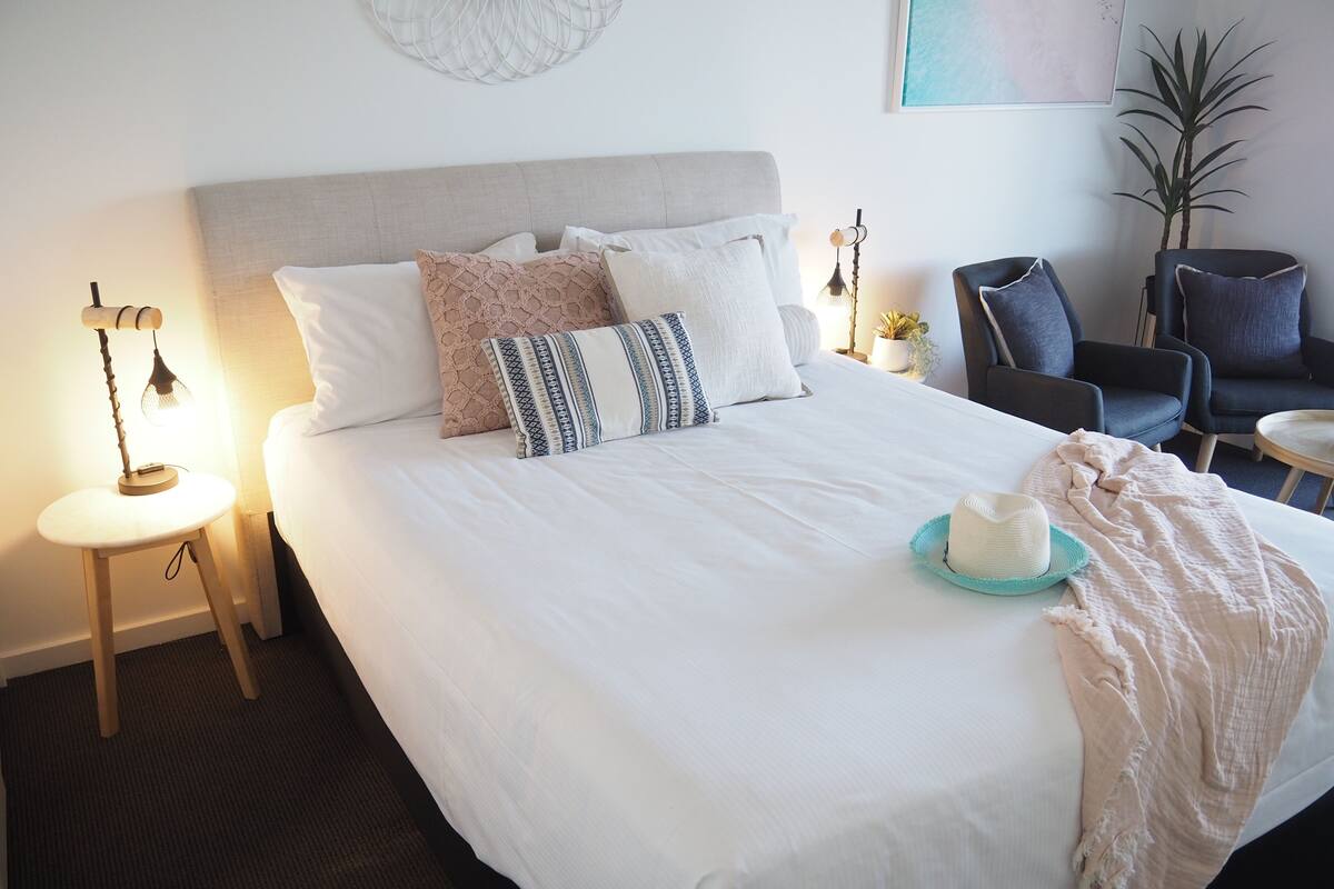 A neatly made bed with a white duvet, multiple pillows, a small round table with a lamp, a chair, and a potted plant.
