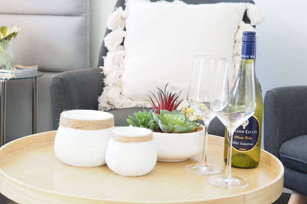 A round wooden table with a bottle of wine, two wine glasses, a potted plant, and two small white containers.
