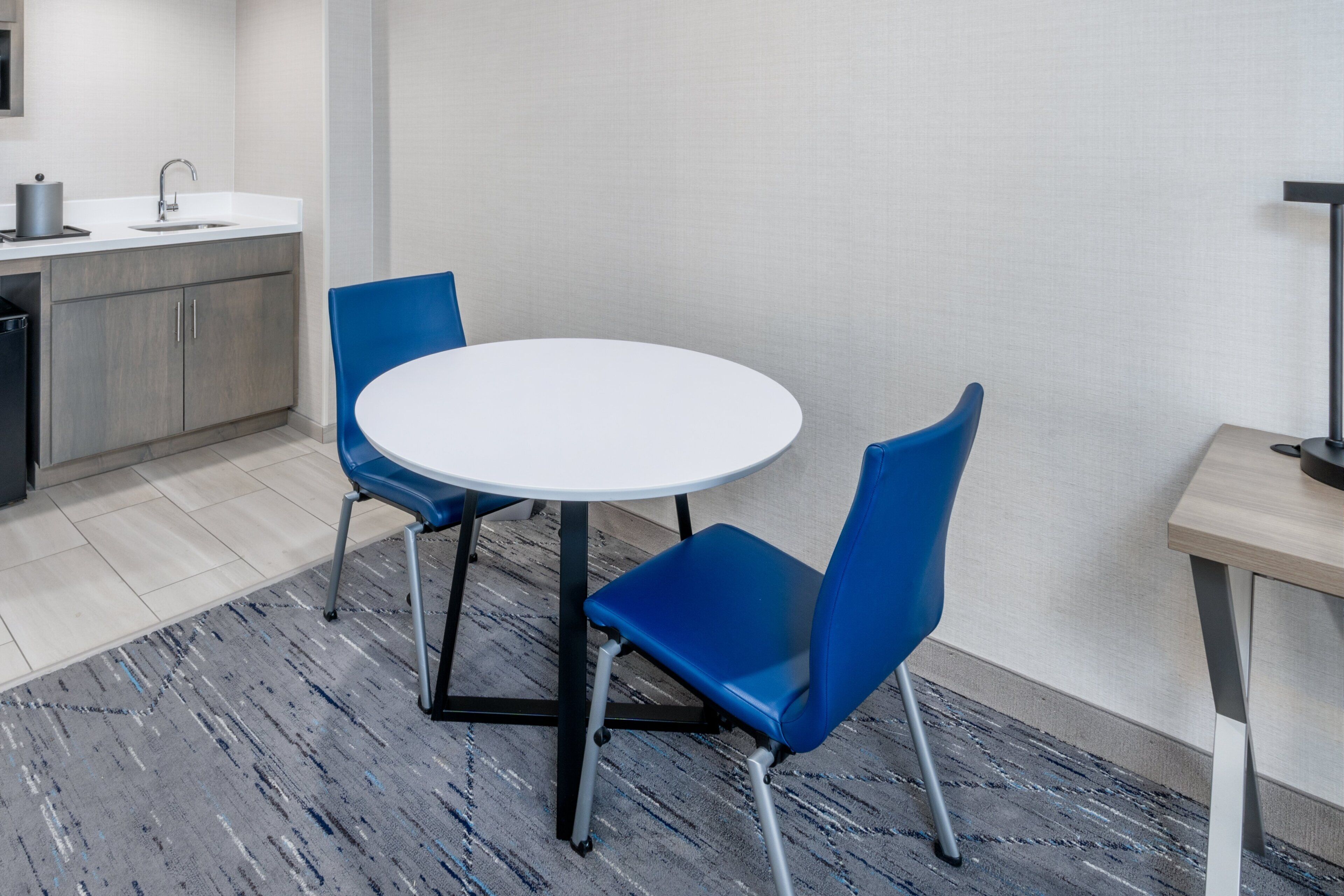 Cocina moderna con encimera blanca, un lavamanos y un mueble pequeño. Hay una mesa redonda con dos sillas azules y una mesa de madera con una lámpara.