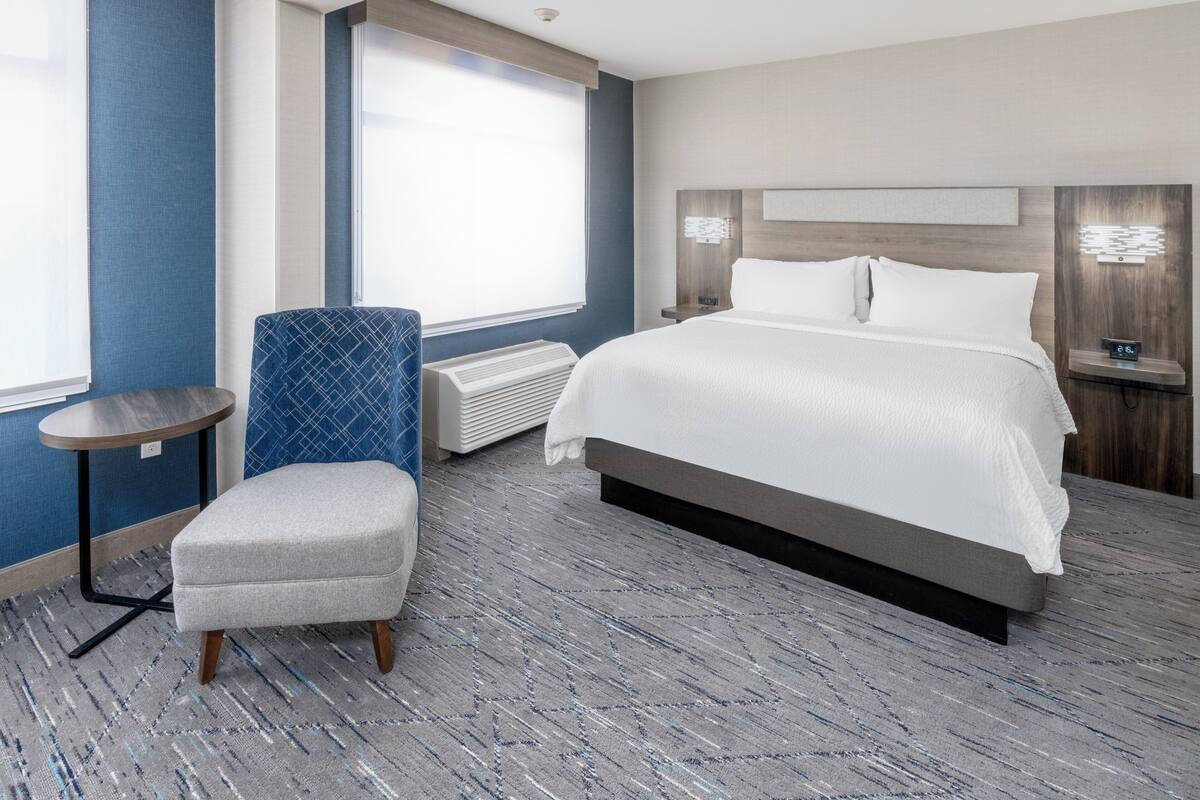 A modern hotel room with a large bed, a seating area, and a window with blinds.