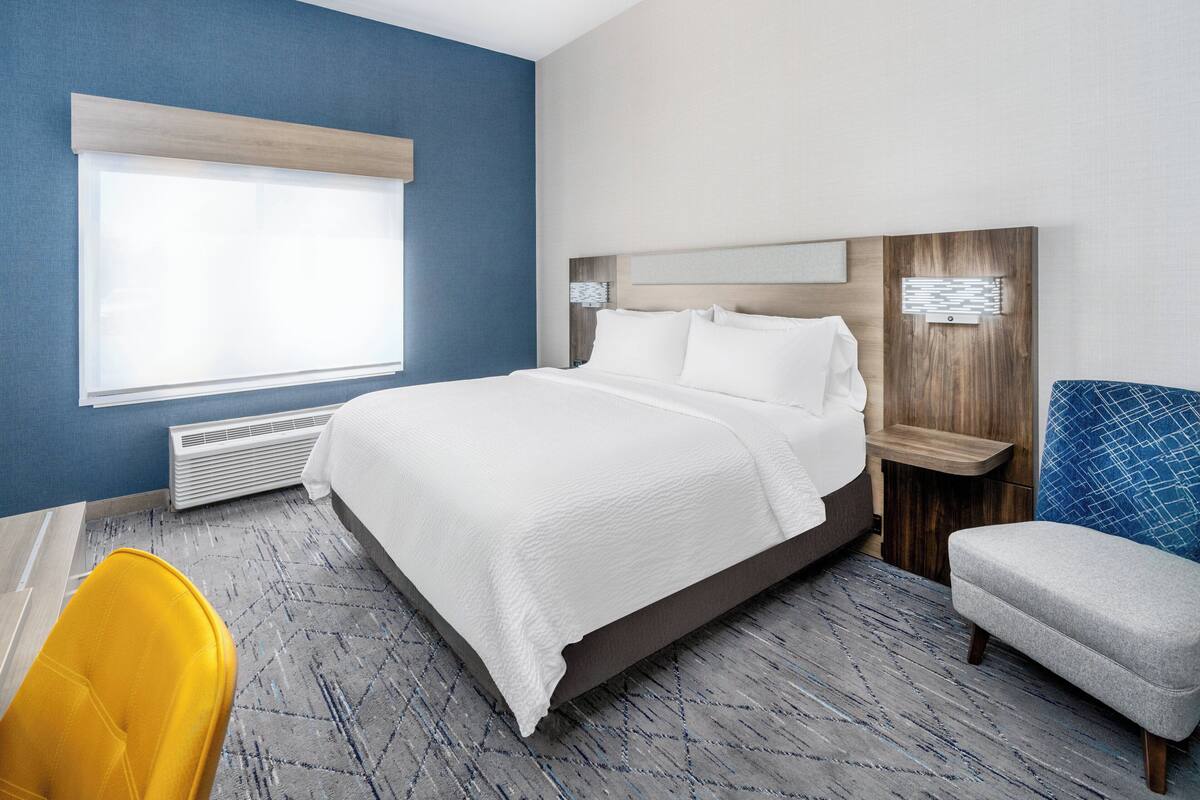 A hotel room with a large bed, a window with blinds, a heater, a yellow chair, and a grey bench.