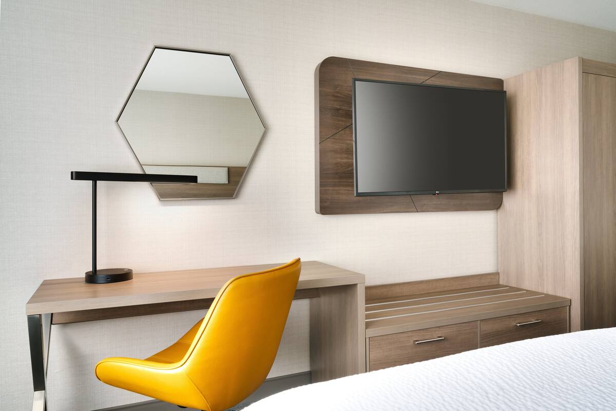 A modern hotel room with a wooden desk, a yellow chair, a flat-screen TV mounted on the wall, and a bed with a white bedspread.