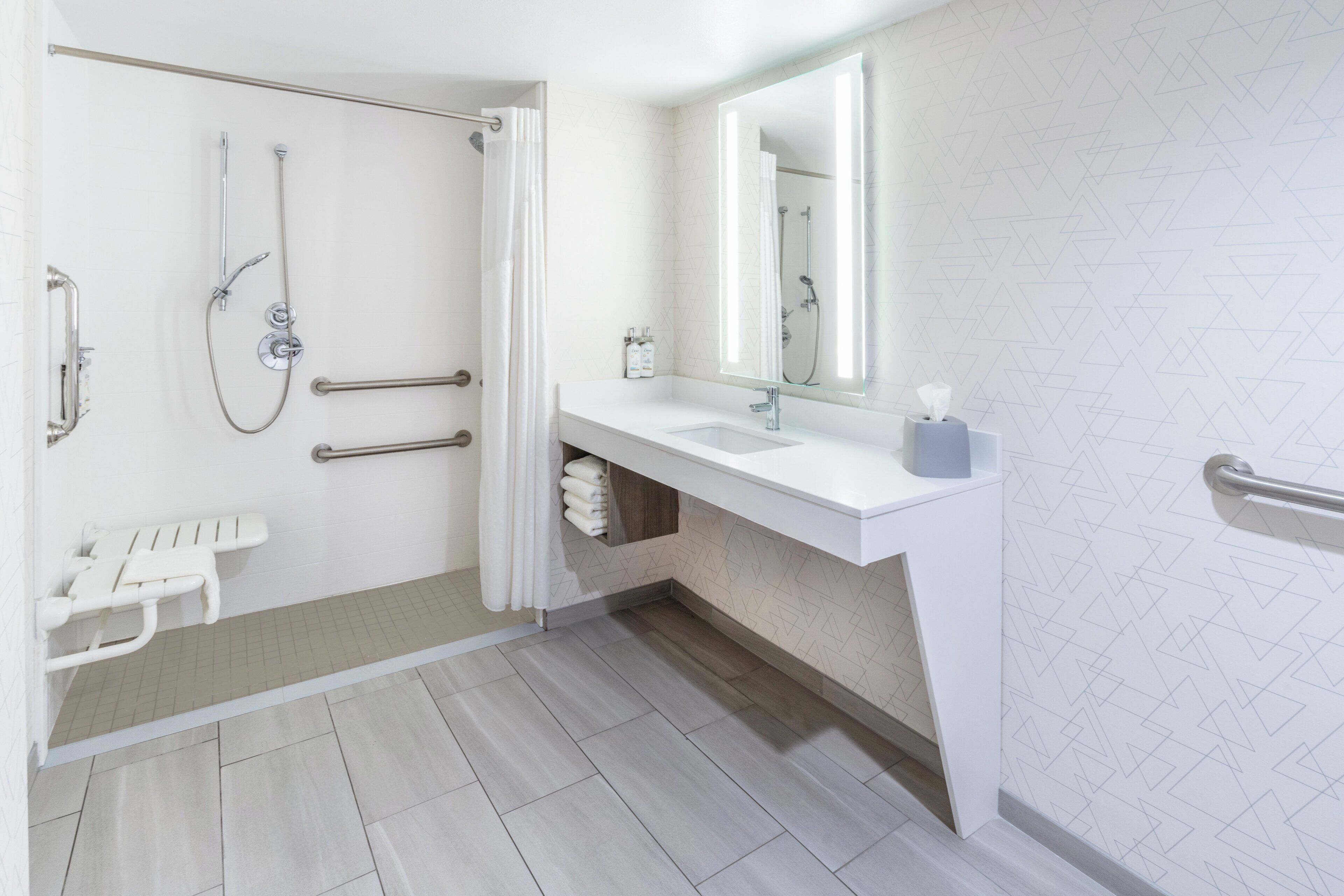 Une salle de bain moderne équipée d’une douche, de barres d’appui, d’un lavabo blanc et d’un grand miroir.