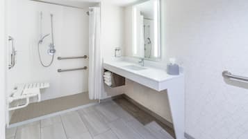 Une salle de bain moderne équipée d’une douche, de barres d’appui, d’un lavabo blanc et d’un grand miroir.
