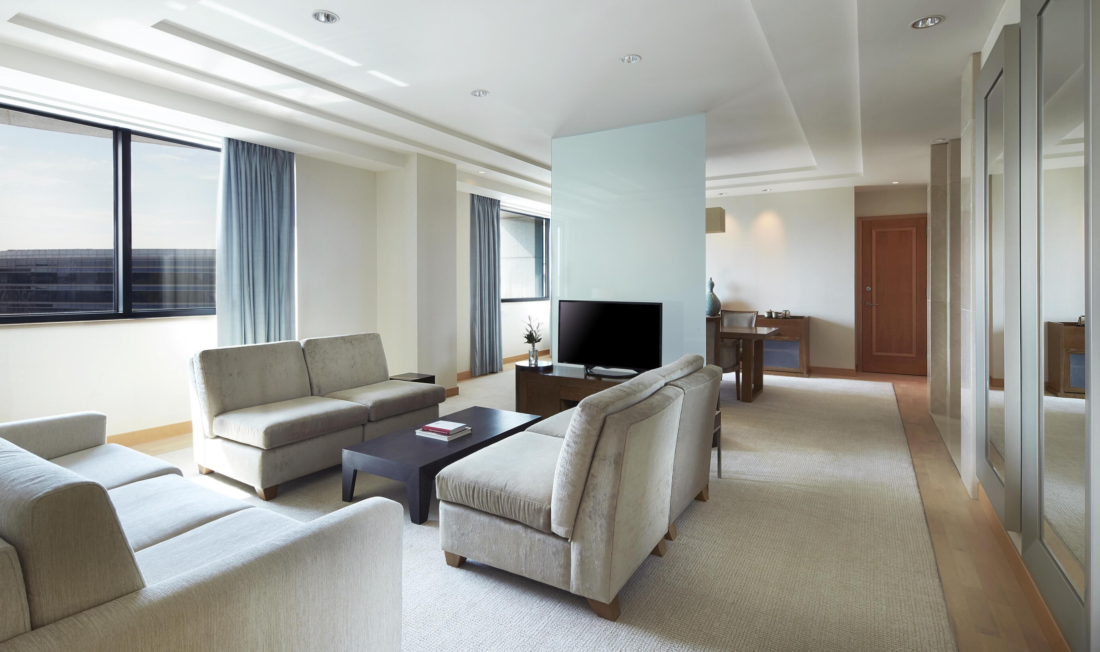A modern hotel room with a sofa, armchairs, a coffee table, a TV, and a small dining area.