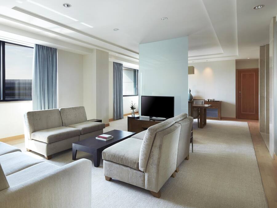 A modern hotel room with a sofa, armchairs, a coffee table, a TV, and a small dining area.