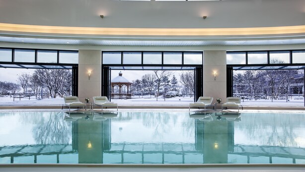 An indoor swimming pool with clear glass walls, lounge chairs, and a view of a snowy outdoor area.