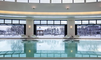 An indoor swimming pool with clear glass walls, lounge chairs, and a view of a snowy outdoor area.