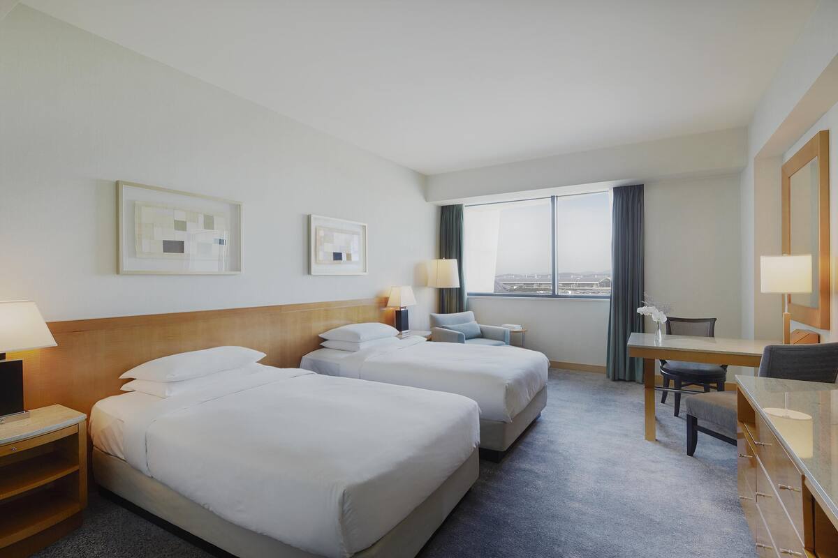 Une chambre d’hôtel avec deux lits, un bureau, une chaise, une lampe et une vue sur la ville.
