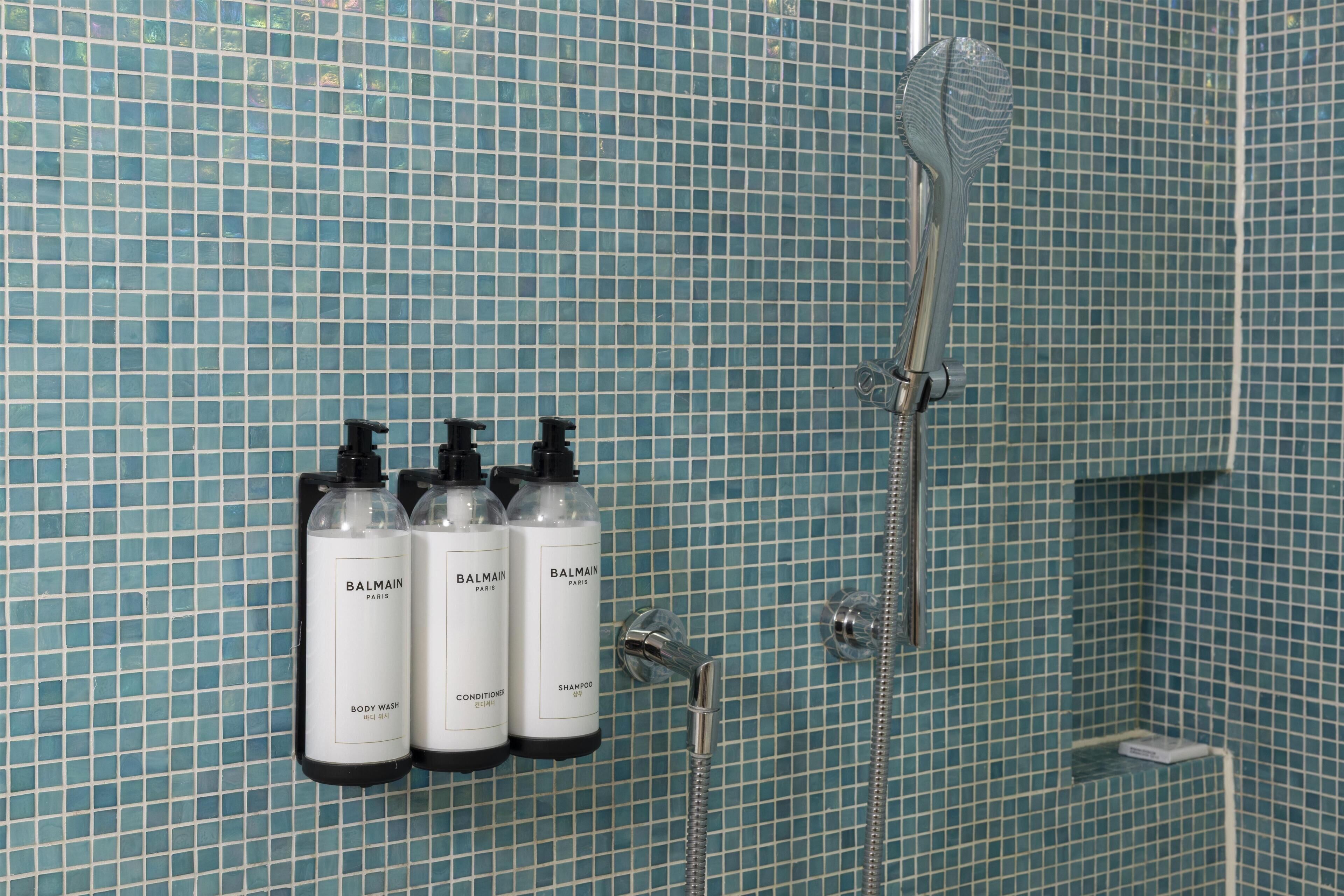A bathroom with a tiled wall, a shower, and three bottles of Balmain Paris body wash, conditioner, and shampoo.