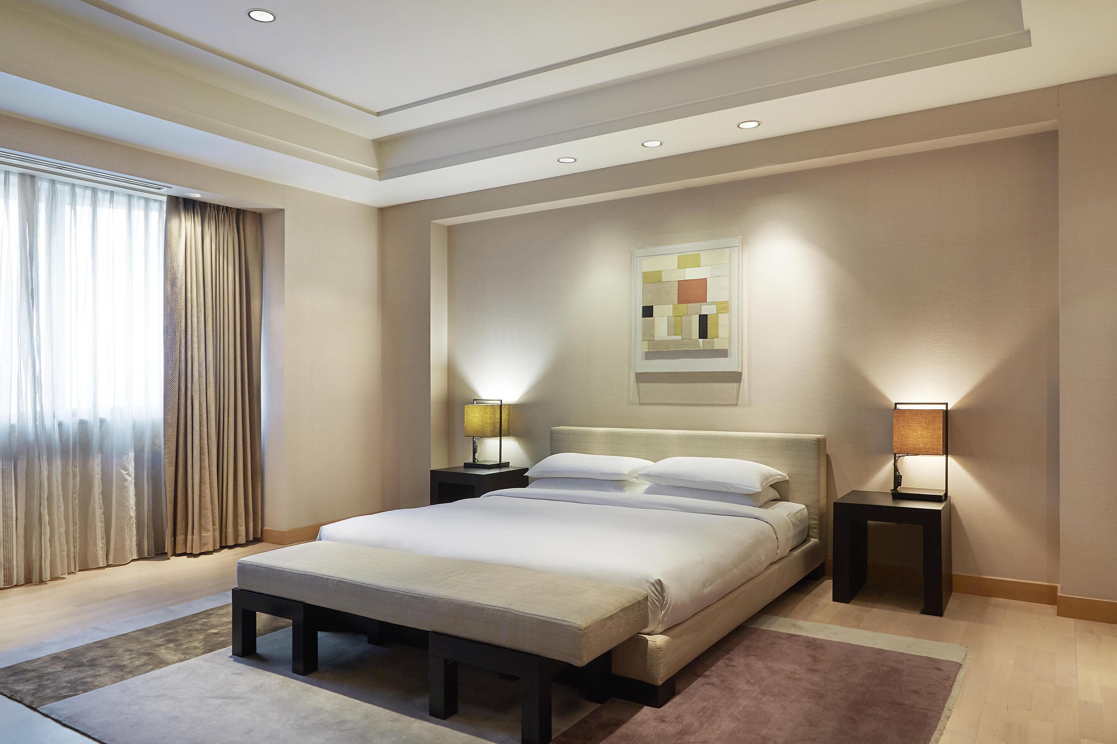 A modern hotel room with a large bed, two bedside tables with lamps, a headboard, and a window with curtains.