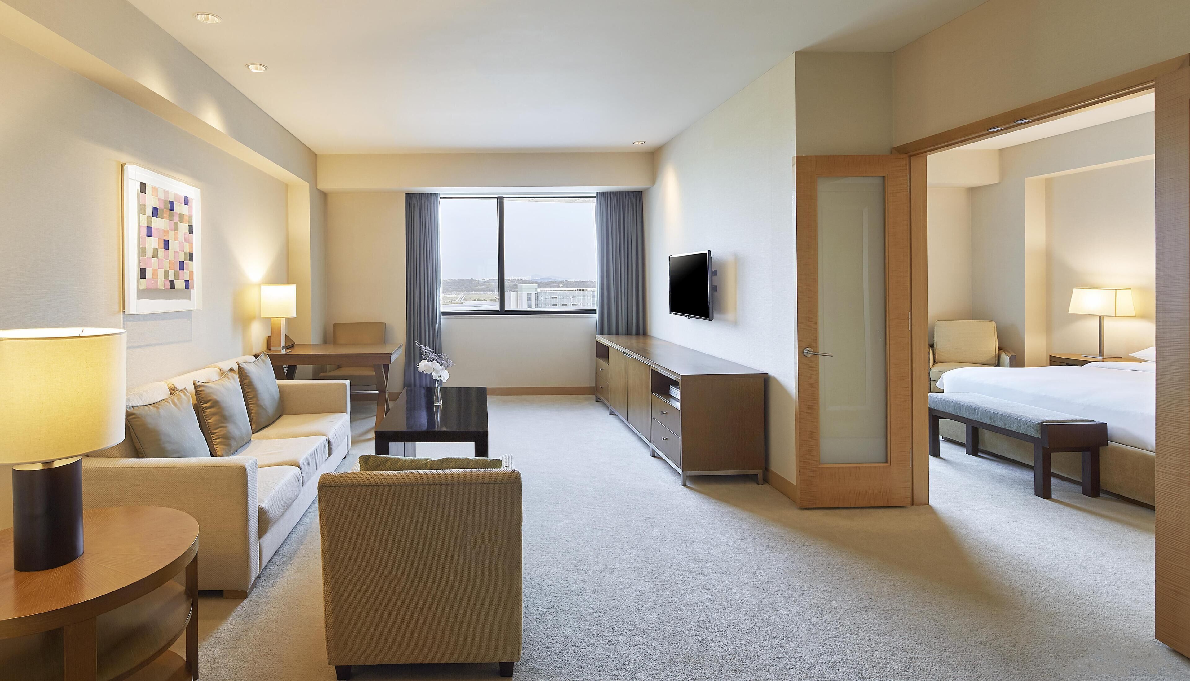 A hotel room with a sofa, a small table, a TV, a bed, and a large window.