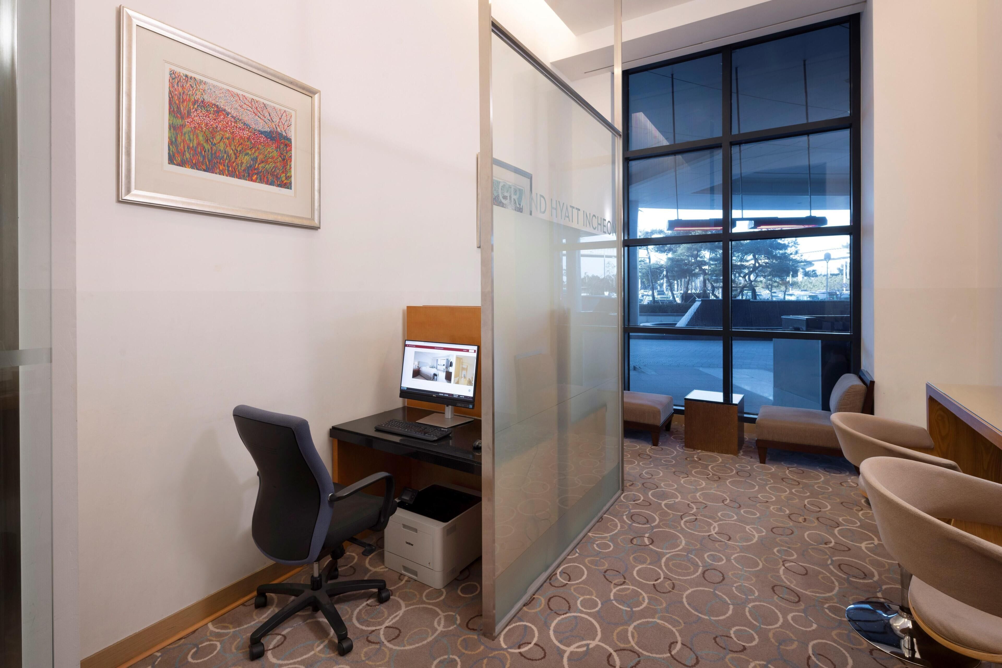 A hotel room with a desk, computer, cushioned seating, a framed picture, and a large window.