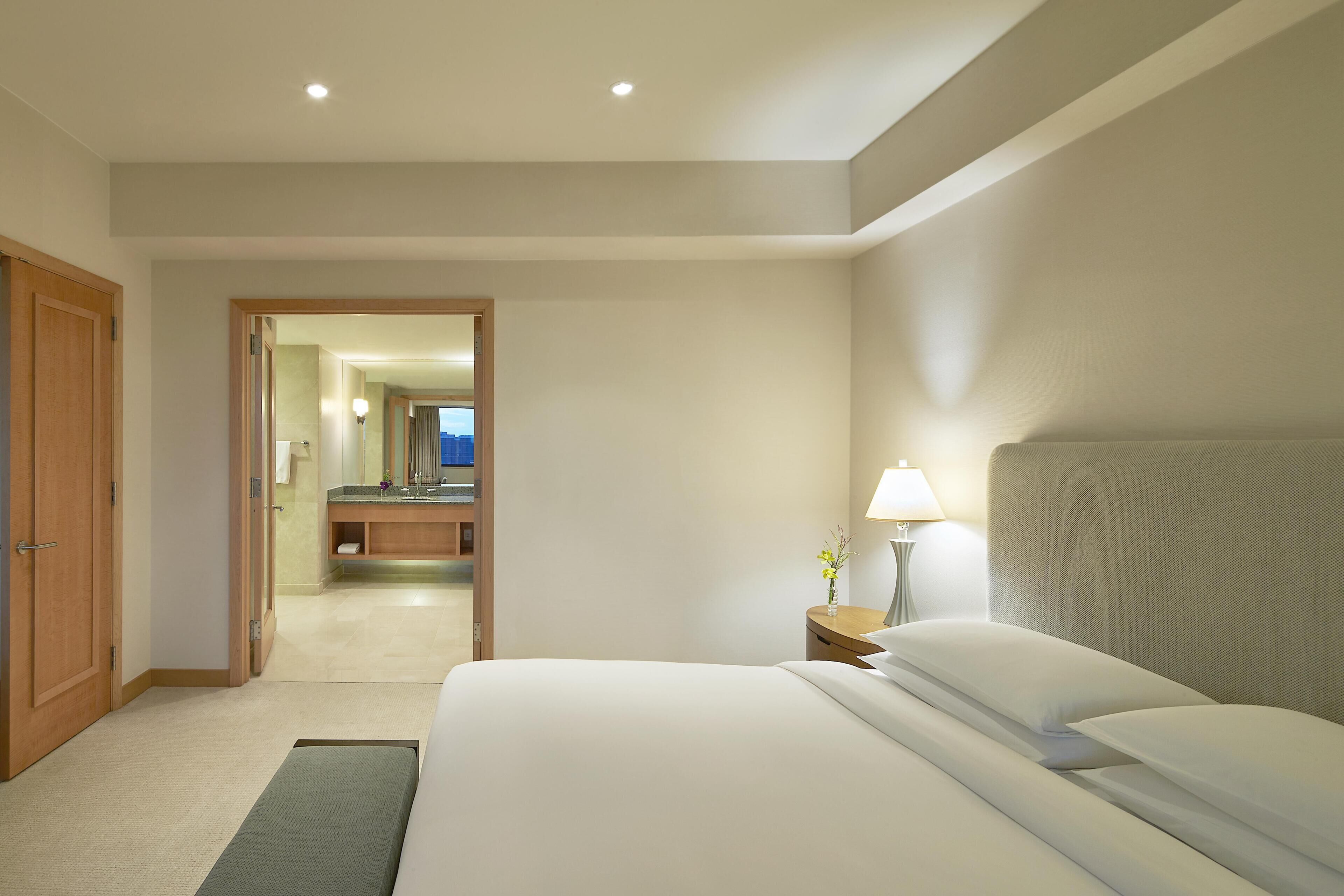 A hotel room with a bed, bedside table, lamp, and a view of the bathroom.