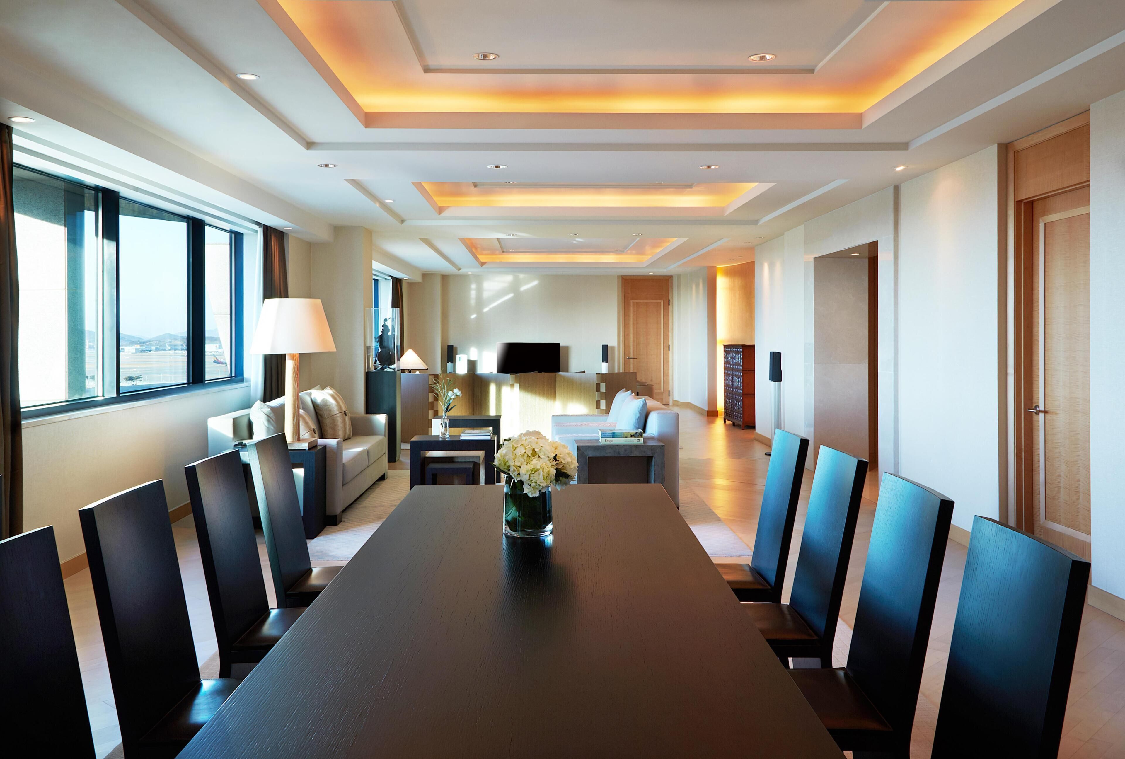 Quarto de hotel com uma grande mesa de jantar, cadeiras, sofá, televisão e vista para a cidade.