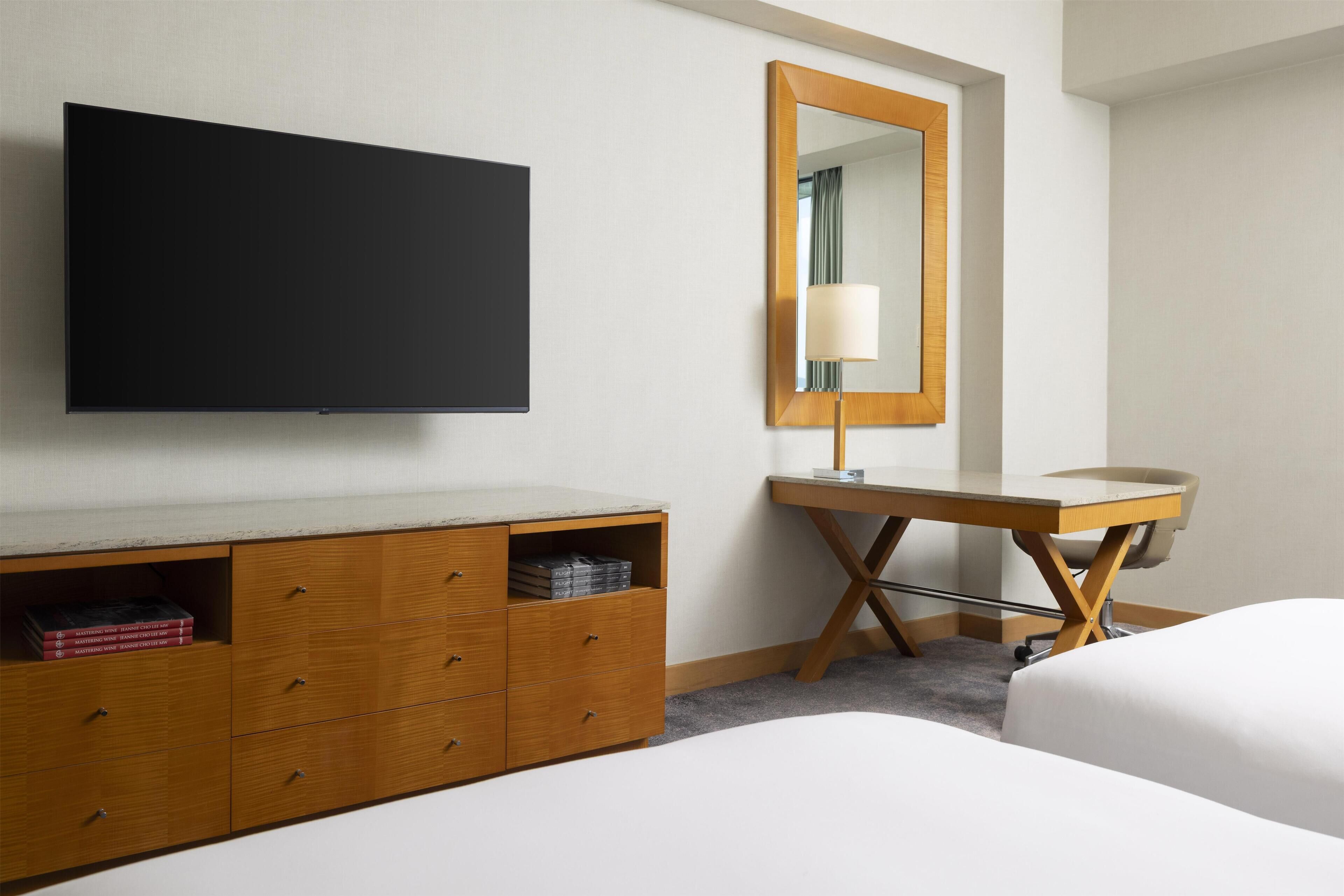 A hotel room with a bed, a wooden dresser, a desk with a chair, a flat-screen TV, and a mirror.